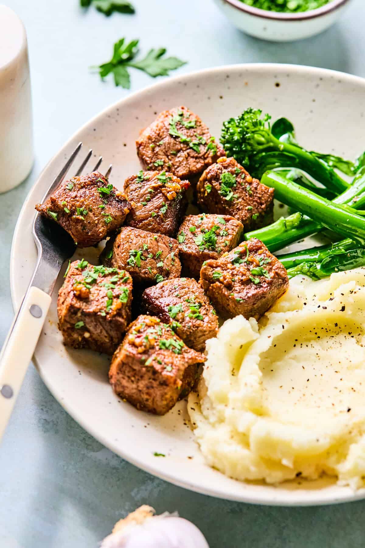 A plate with garlic steak bites, mashed potatoes with black pepper, and steamed broccolini, garnished with chopped herbs. A fork rests on the plate’s edge.