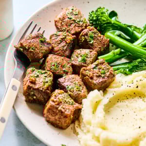 A plate with garlic steak bites, mashed potatoes with black pepper, and steamed broccolini, garnished with chopped herbs. A fork rests on the plate’s edge.