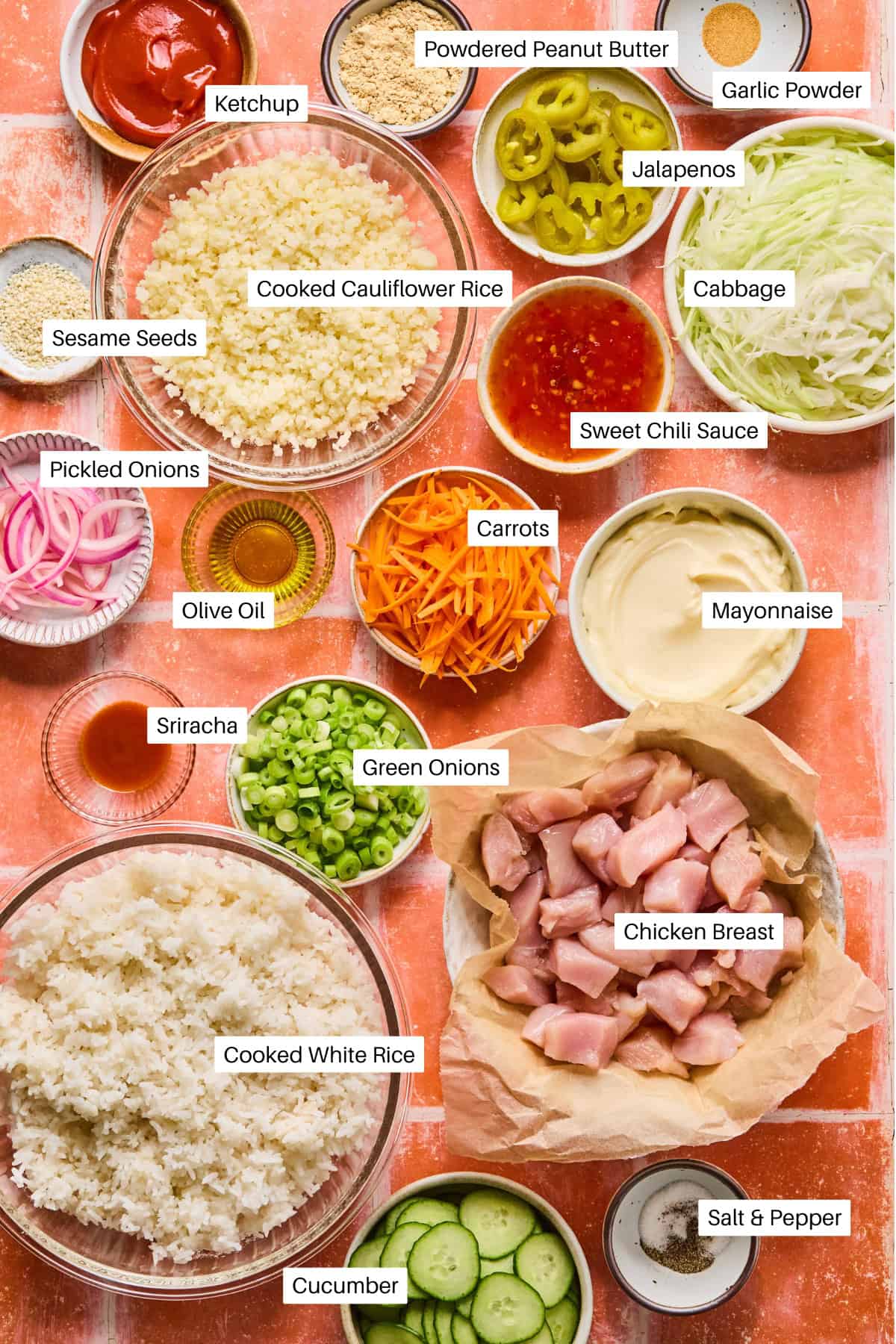 An assortment of labeled ingredients on a table for bang bang chicken, including chicken breast, cooked white rice, cauliflower rice, carrots, cabbage, green onions, cucumber, jalapeños, mayonnaise, sriracha, sweet chili sauce, pickled onions, and seasonings.