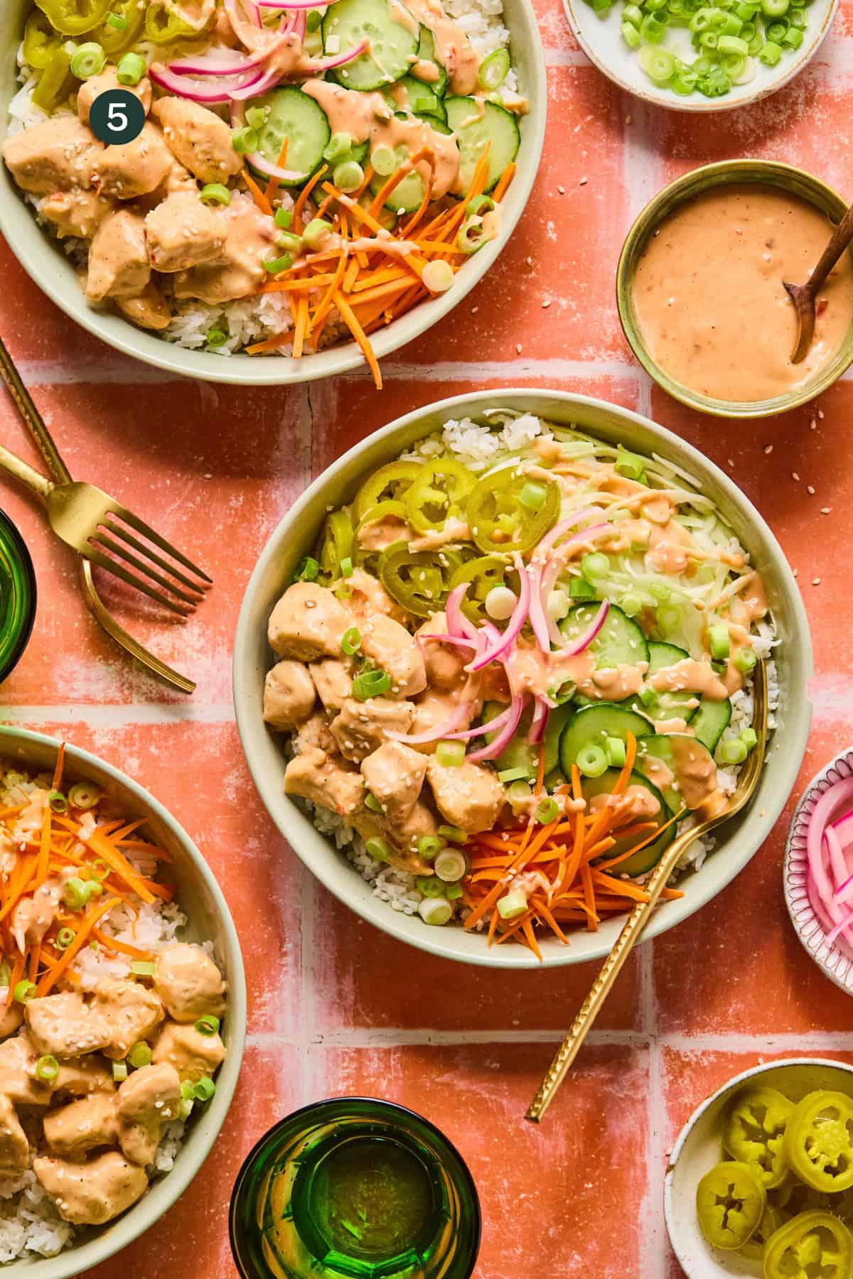 A top-down view of bang bang chicken rice bowls topped with sliced chicken, shredded carrots, pickled onions, cucumbers, jalapeños, green onions, and creamy sauce, surrounded by forks, green glasses, and side dishes.