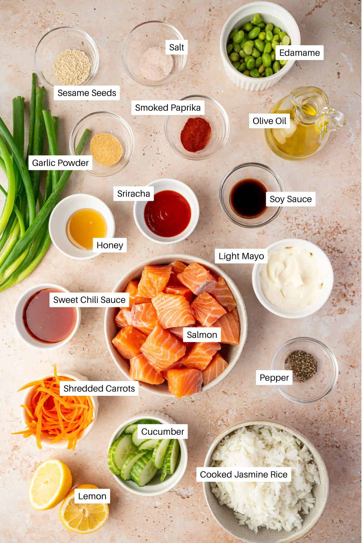 Bowls and containers of poke bowl ingredients, including bang bang salmon, jasmine rice, cucumber, shredded carrots, lemon, sauces, seasonings, edamame, olive oil, and green onions arranged on a light surface.