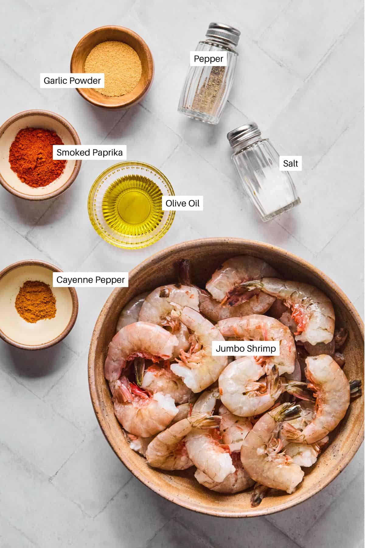 A bowl of raw jumbo shrimp, perfect for Blackened Shrimp, surrounded by small dishes of garlic powder, smoked paprika, cayenne pepper, olive oil, and salt and pepper shakers—all labeled on a light-colored surface.
