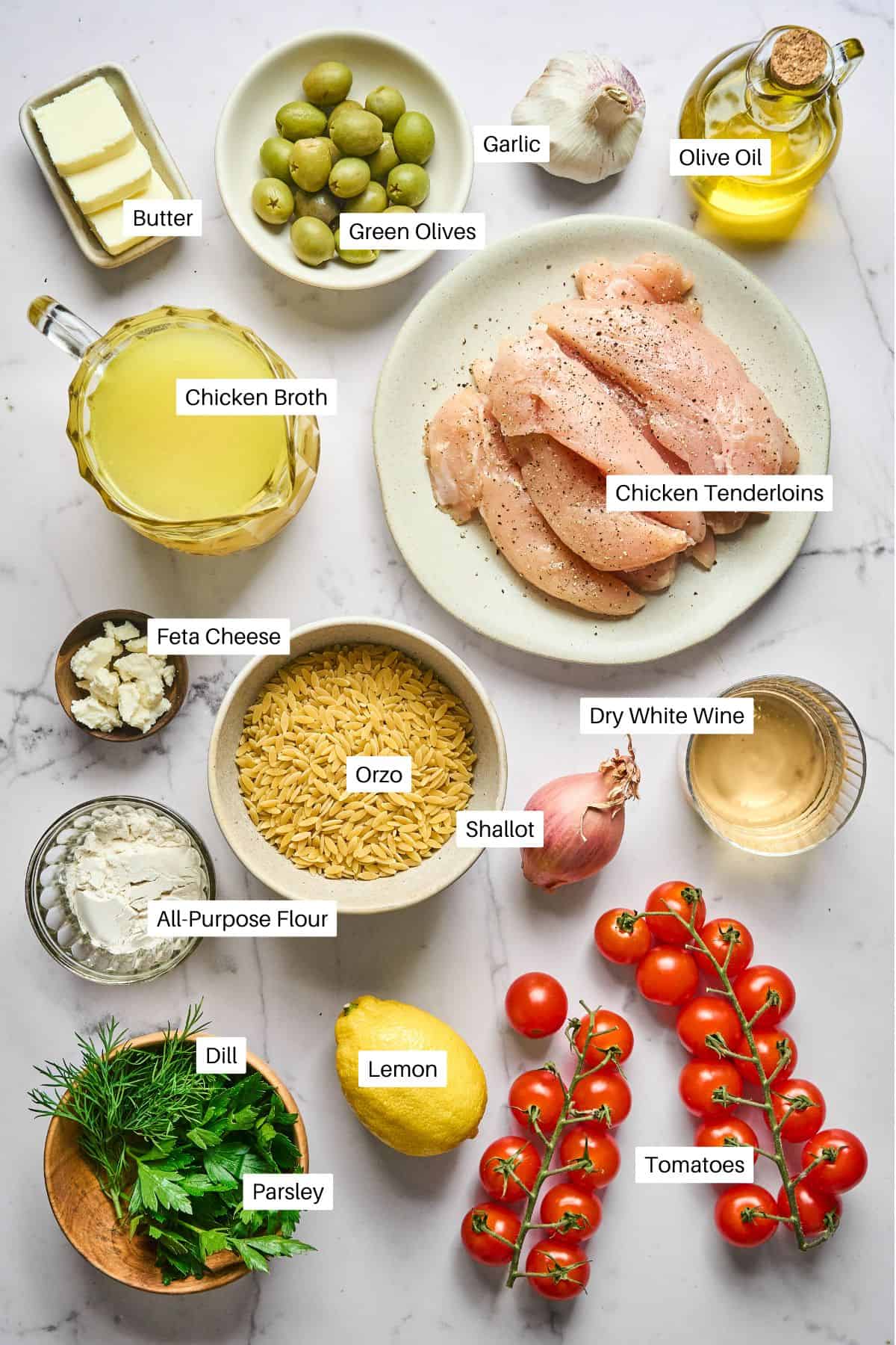 A top-down view of labeled ingredients for lemon orzo chicken pasta on a white surface: chicken tenderloins, green olives, garlic, olive oil, butter, chicken broth, feta cheese, orzo, dry white wine, shallot, flour, dill, parsley, lemon, and tomatoes.