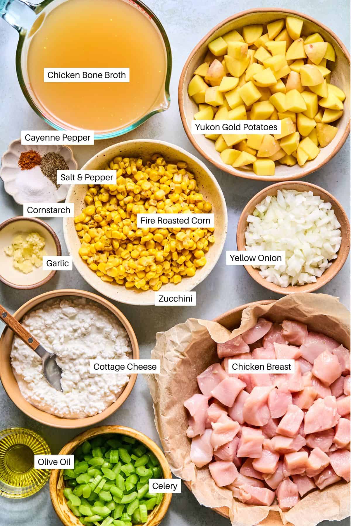 Assorted labeled ingredients for a hearty Corn and Potato Chowder are arranged in bowls, including chicken breast, Yukon gold potatoes, corn, onion, zucchini, celery, garlic, cottage cheese, olive oil, chicken bone broth, and seasonings.