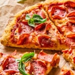 Close-up of sliced cottage cheese pizza with melted cheese, crispy crust, and fresh basil leaves on top, displayed on parchment paper with herbs and pizza toppings in the background.