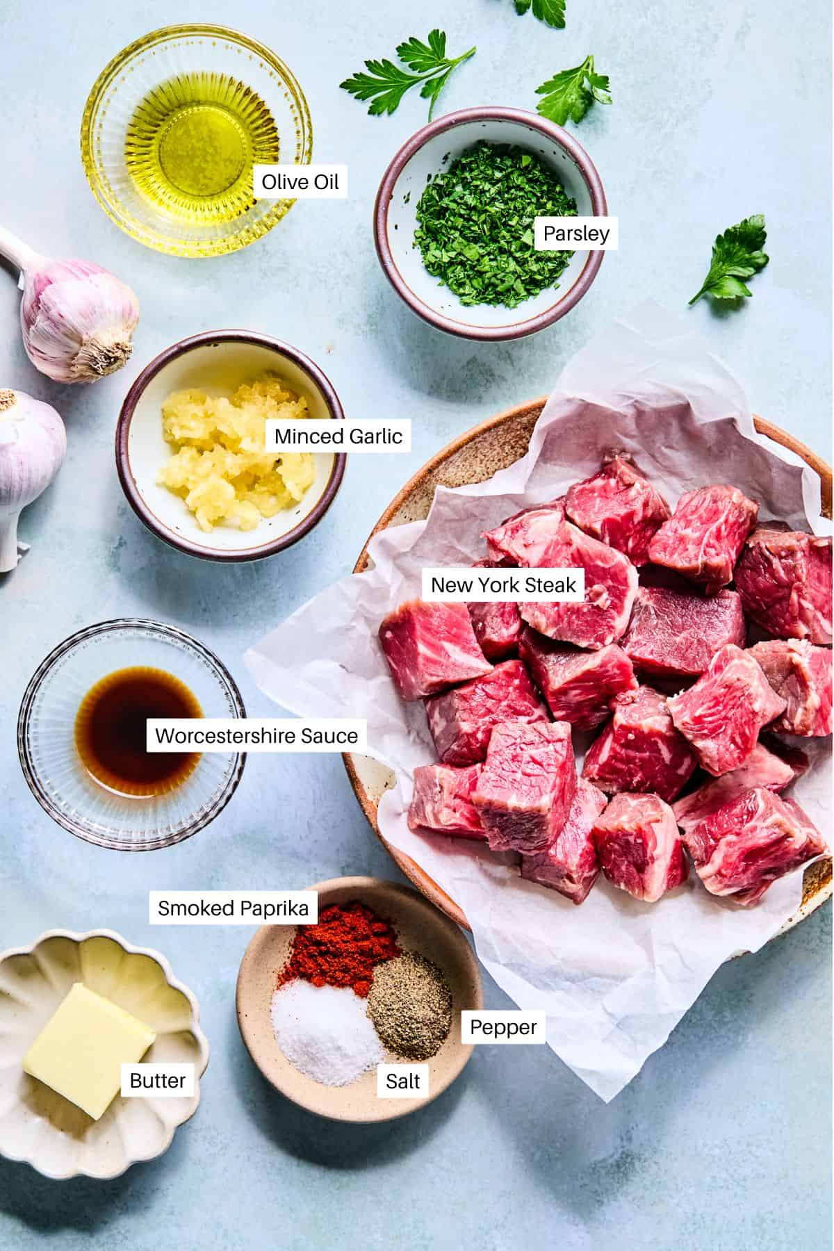 Overhead view of labeled ingredients for garlic steak bites, featuring cubed New York steak, parsley, olive oil, minced garlic, Worcestershire sauce, smoked paprika, salt, pepper, and a pat of butter on a light background.
