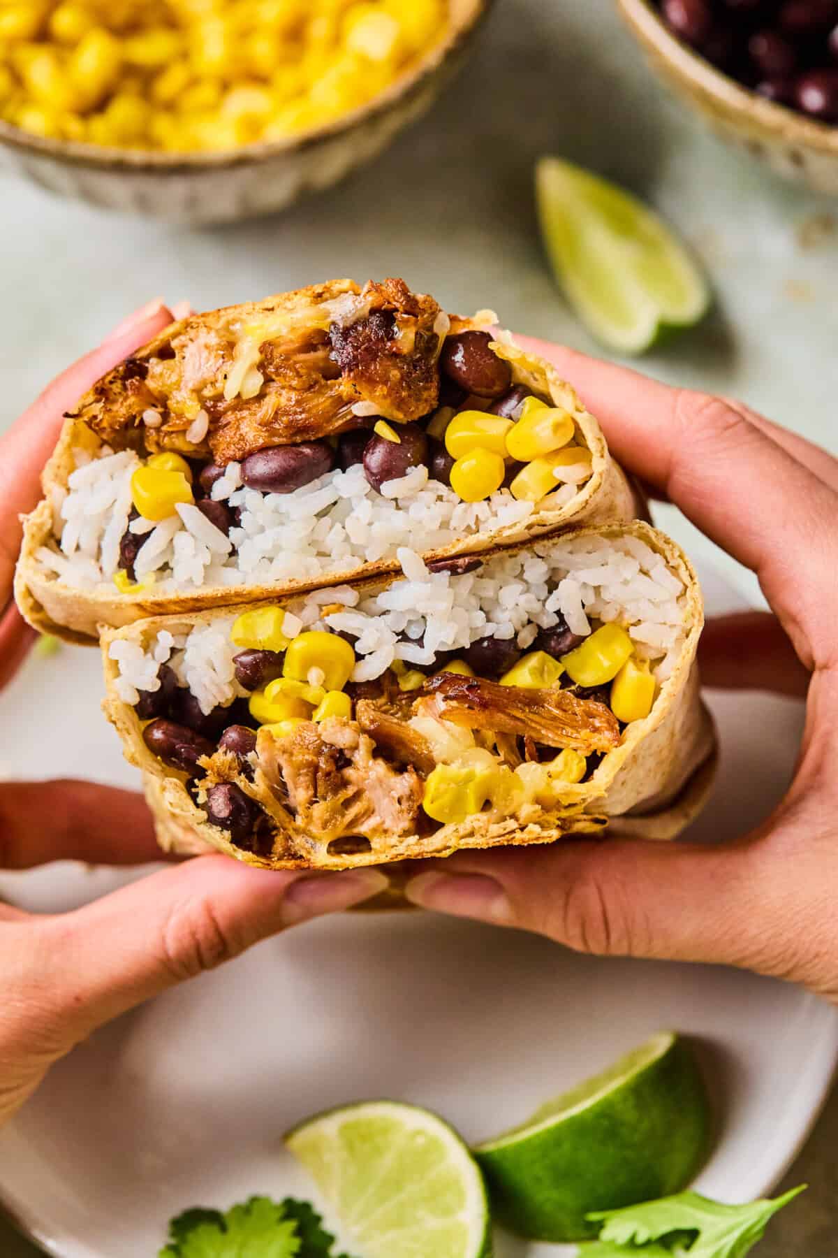 Two hands hold a Carnitas Burrito cut in half, revealing rice, black beans, corn, and shredded meat. Lime wedges, cilantro, and bowls of corn and beans are visible in the background.