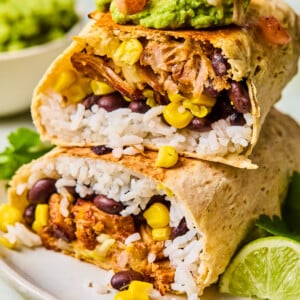 A plate with two Carnitas Burritos cut in half, filled with rice, black beans, corn, and shredded meat, topped with guacamole and salsa. Lime wedges and cilantro are on the side, with extra guacamole in the background.