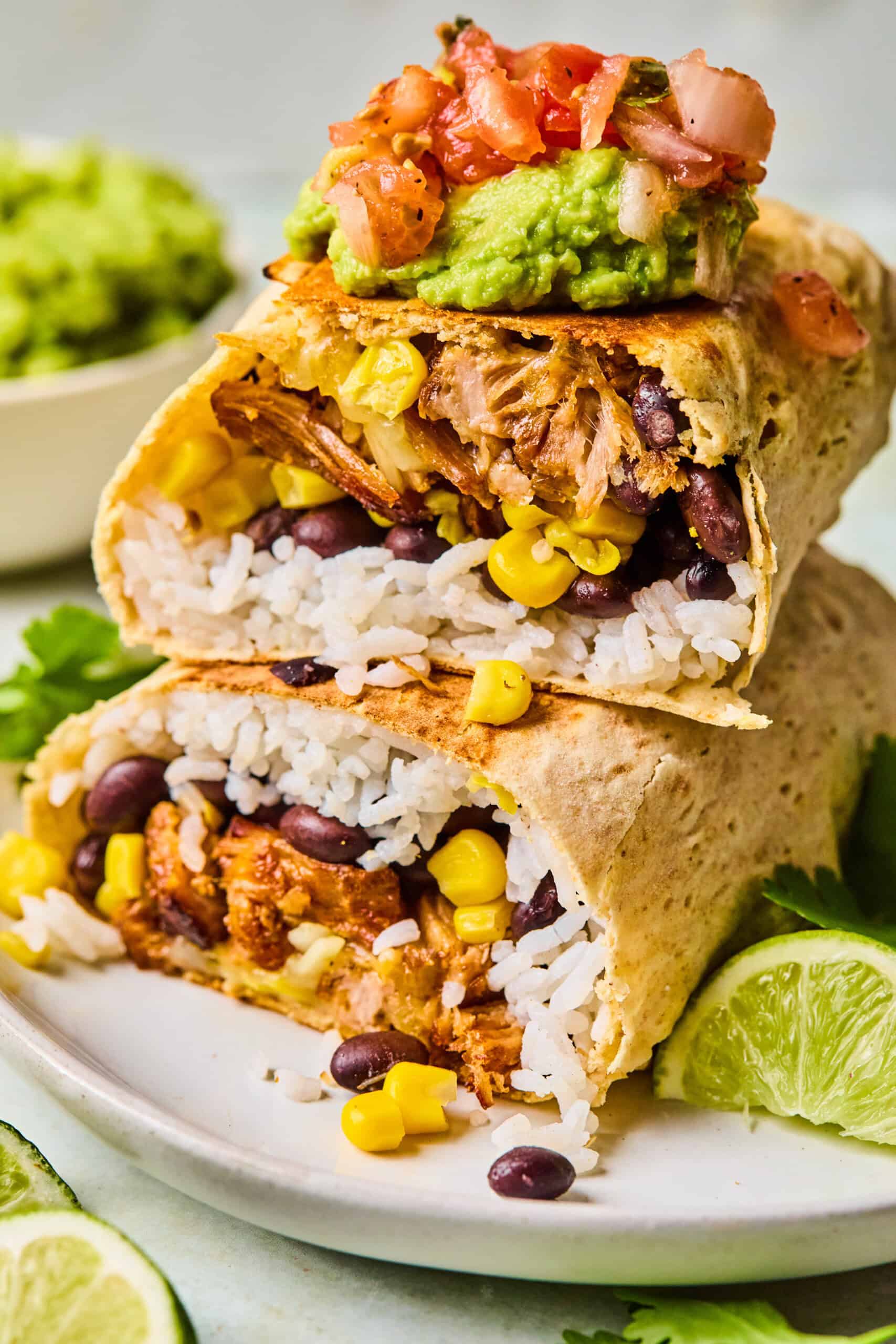 A plate with two Carnitas Burritos cut in half, filled with rice, black beans, corn, and shredded meat, topped with guacamole and salsa. Lime wedges and cilantro are on the side, with extra guacamole in the background.