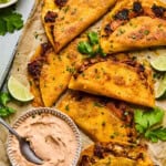 A tray of crispy Instant Pot Carnitas Tacos filled with juicy meat is garnished with fresh cilantro and lime wedges. A bowl of creamy dipping sauce with a spoon and a bowl of guacamole sit nearby, all arranged on parchment paper.