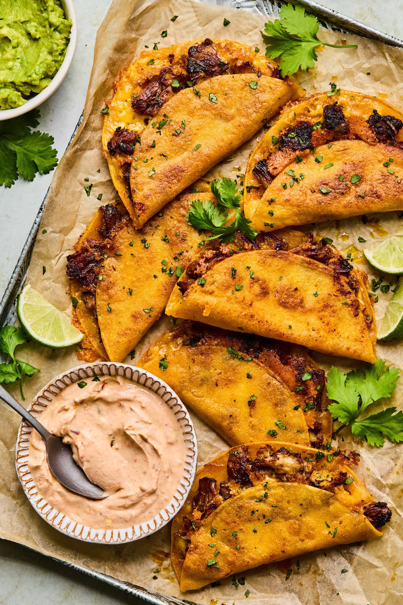 A tray of crispy Instant Pot Carnitas Tacos filled with juicy meat is garnished with fresh cilantro and lime wedges. A bowl of creamy dipping sauce with a spoon and a bowl of guacamole sit nearby, all arranged on parchment paper.