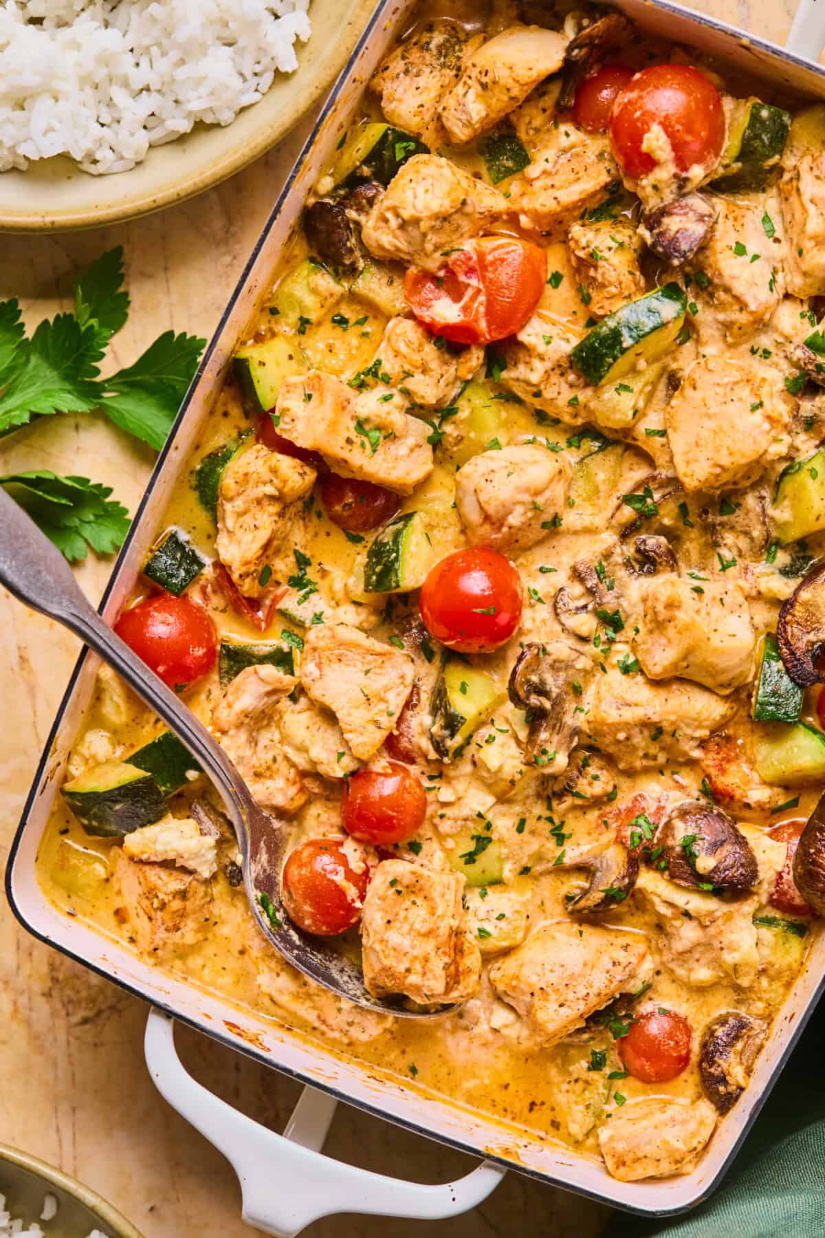 A close-up of a creamy chicken feta recipe in a white baking dish, featuring chunks of chicken, cherry tomatoes, zucchini, and mushrooms, garnished with herbs. A serving spoon rests inside, with rice and parsley on the side.