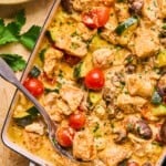 A close-up of a creamy chicken feta recipe in a white baking dish, featuring chunks of chicken, cherry tomatoes, zucchini, and mushrooms, garnished with herbs. A serving spoon rests inside, with rice and parsley on the side.