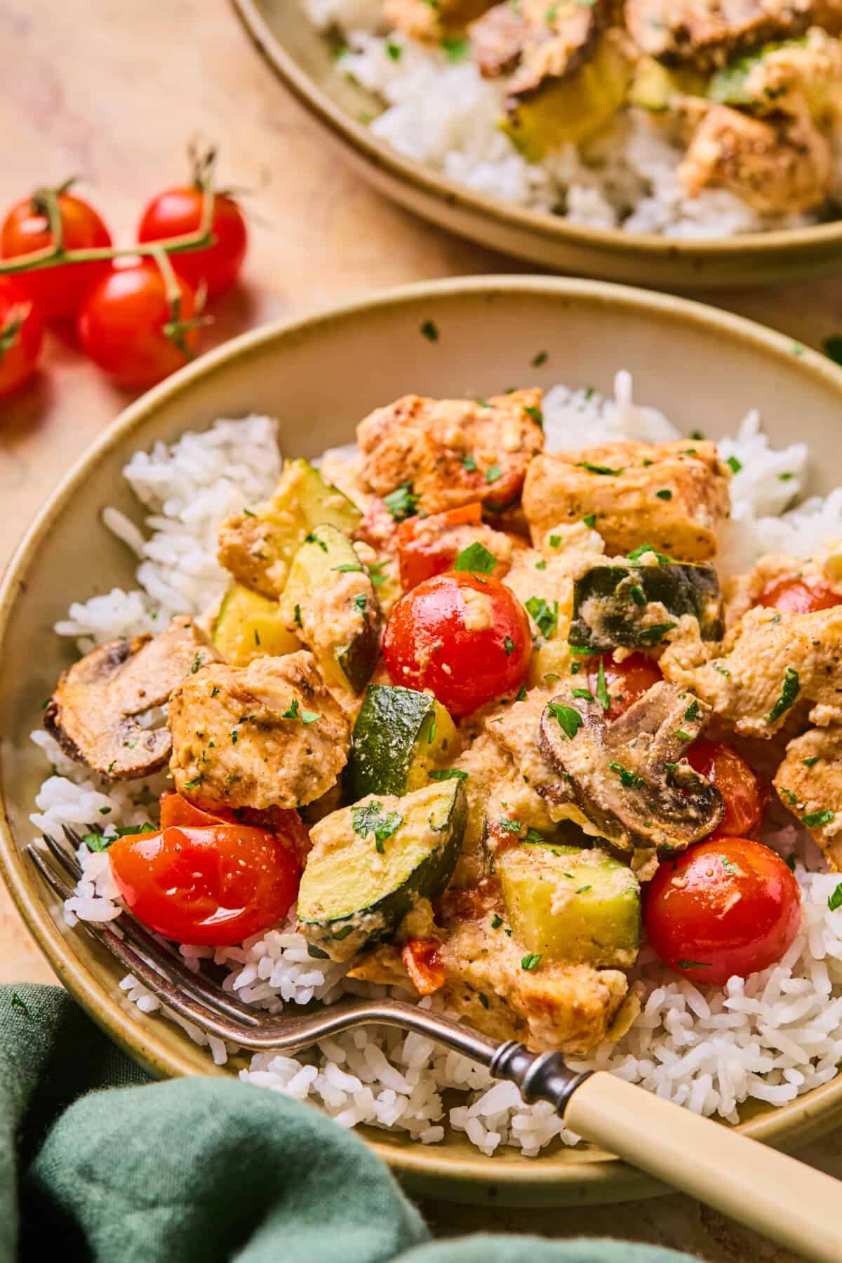 A bowl of white rice topped with creamy chicken, zucchini, mushrooms, and cherry tomatoes, garnished with chopped herbs. Inspired by a chicken feta recipe, a fork rests in the bowl, and fresh tomatoes are nearby.