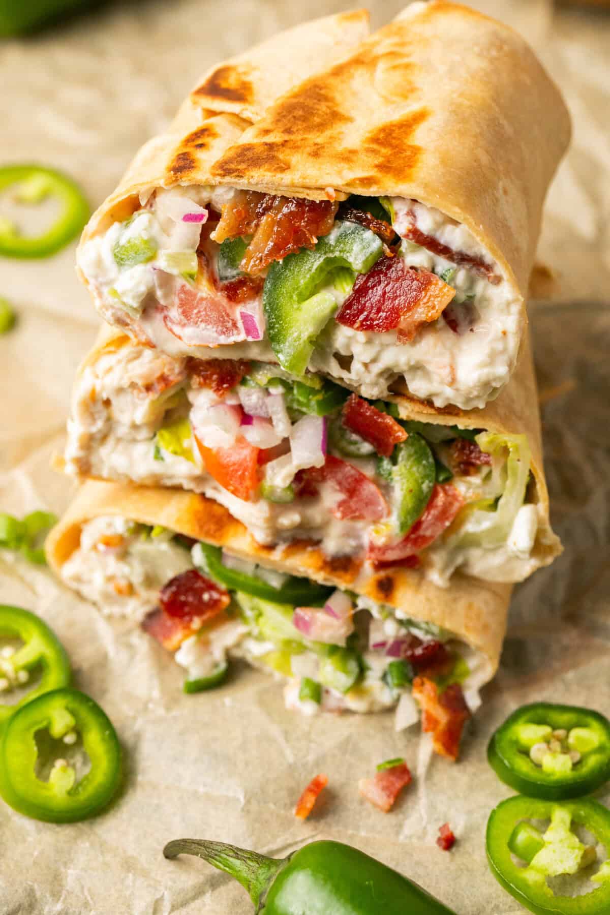 Three grilled shredded chicken wraps are stacked, filled with cream cheese, chopped tomatoes, bacon, red onions, and jalapeño slices. Sliced jalapeños are scattered around on parchment paper.
