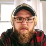 A bearded man wearing glasses, a dark baseball cap, and large over-ear headphones sits indoors, looking directly at the camera. He is dressed in a red and black plaid jacket, possibly checking the oh snap macros app. Bright windows are in the background.
