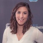 A woman with medium-length brown hair wearing a white blouse smiles at the camera against a gray background with faint text and logo elements, reflecting the approachable vibe of the oh snap macros app.