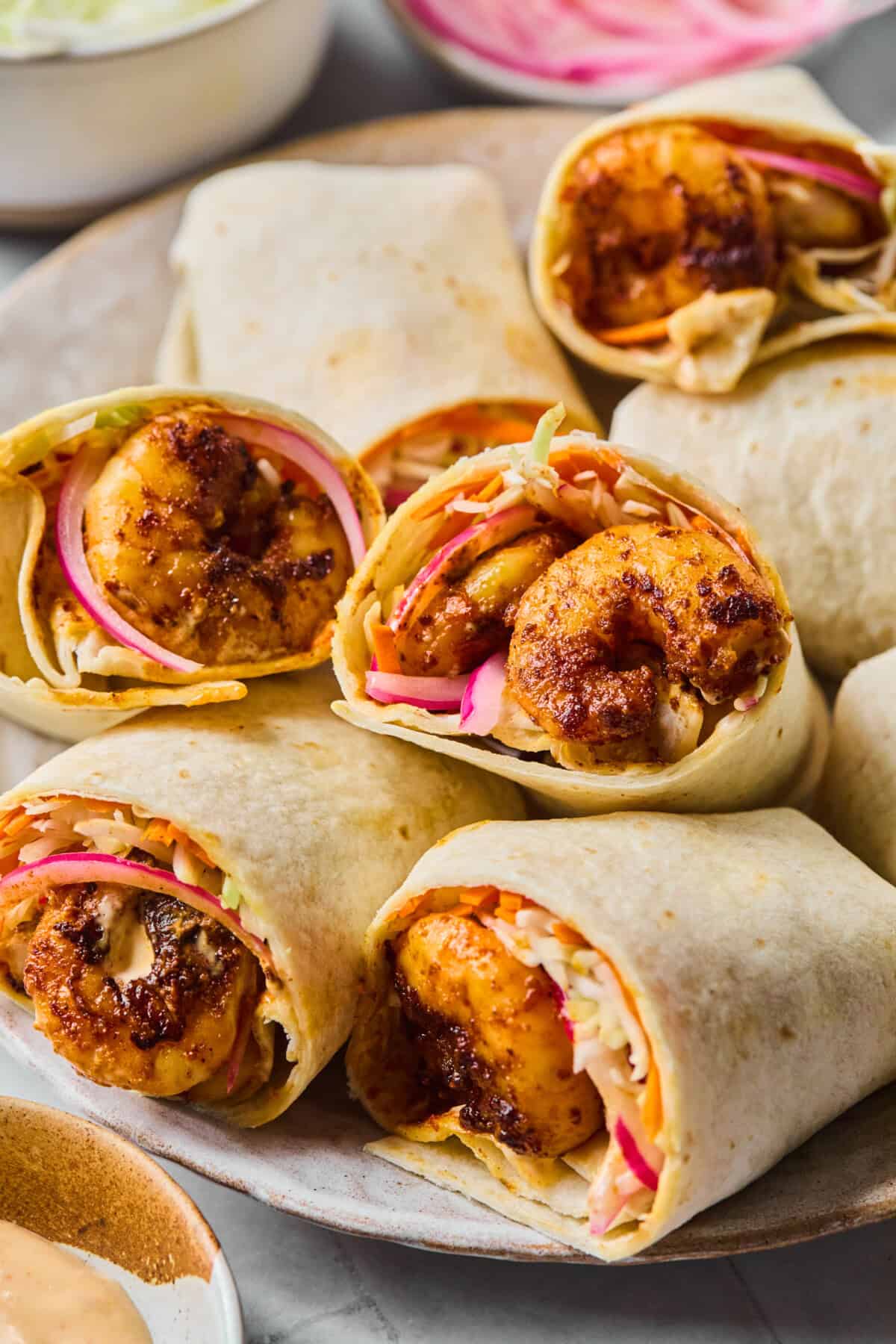A plate of Shrimp Wraps in flour tortillas, filled with seasoned shrimp, sliced red onions, and shredded cabbage. The wraps are arranged closely together, some cut in half to reveal the vibrant filling.