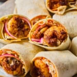 A plate of Shrimp Wraps in flour tortillas, filled with seasoned shrimp, sliced red onions, and shredded cabbage. The wraps are arranged closely together, some cut in half to reveal the vibrant filling.