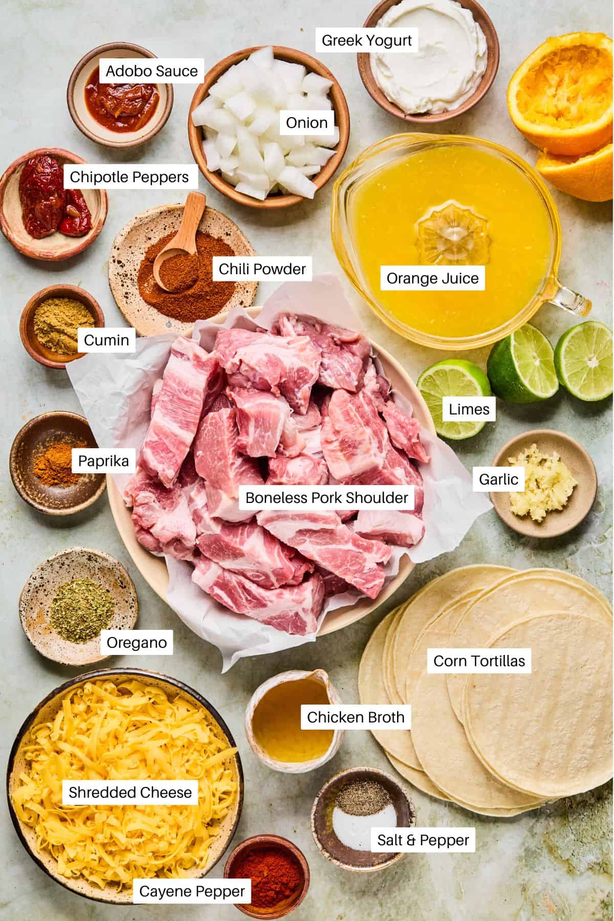 Overhead view of labeled ingredients for Instant Pot Carnitas Tacos, including boneless pork shoulder, shredded cheese, corn tortillas, orange juice, onion, garlic, spices, chipotle peppers in adobo sauce, limes, Greek yogurt, chicken broth, salt & pepper.