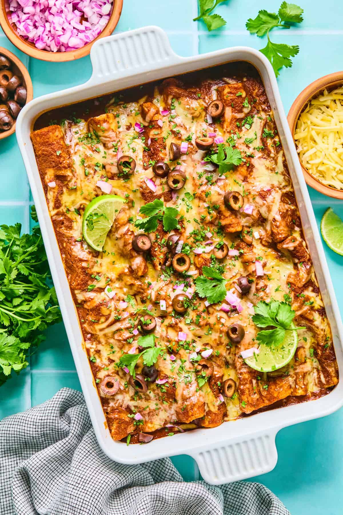 A baking dish filled with cheesy rotisserie chicken enchiladas, topped with sliced black olives, red onions, cilantro, and lime wedges. Surrounding the dish are small bowls of chopped onions, shredded cheese, cilantro, and a gray cloth.