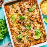 A baking dish filled with cheesy rotisserie chicken enchiladas, topped with sliced black olives, red onions, cilantro, and lime wedges. Surrounding the dish are small bowls of chopped onions, shredded cheese, cilantro, and a gray cloth.