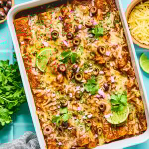 A baking dish filled with cheesy rotisserie chicken enchiladas, topped with sliced black olives, red onions, cilantro, and lime wedges. Surrounding the dish are small bowls of chopped onions, shredded cheese, cilantro, and a gray cloth.