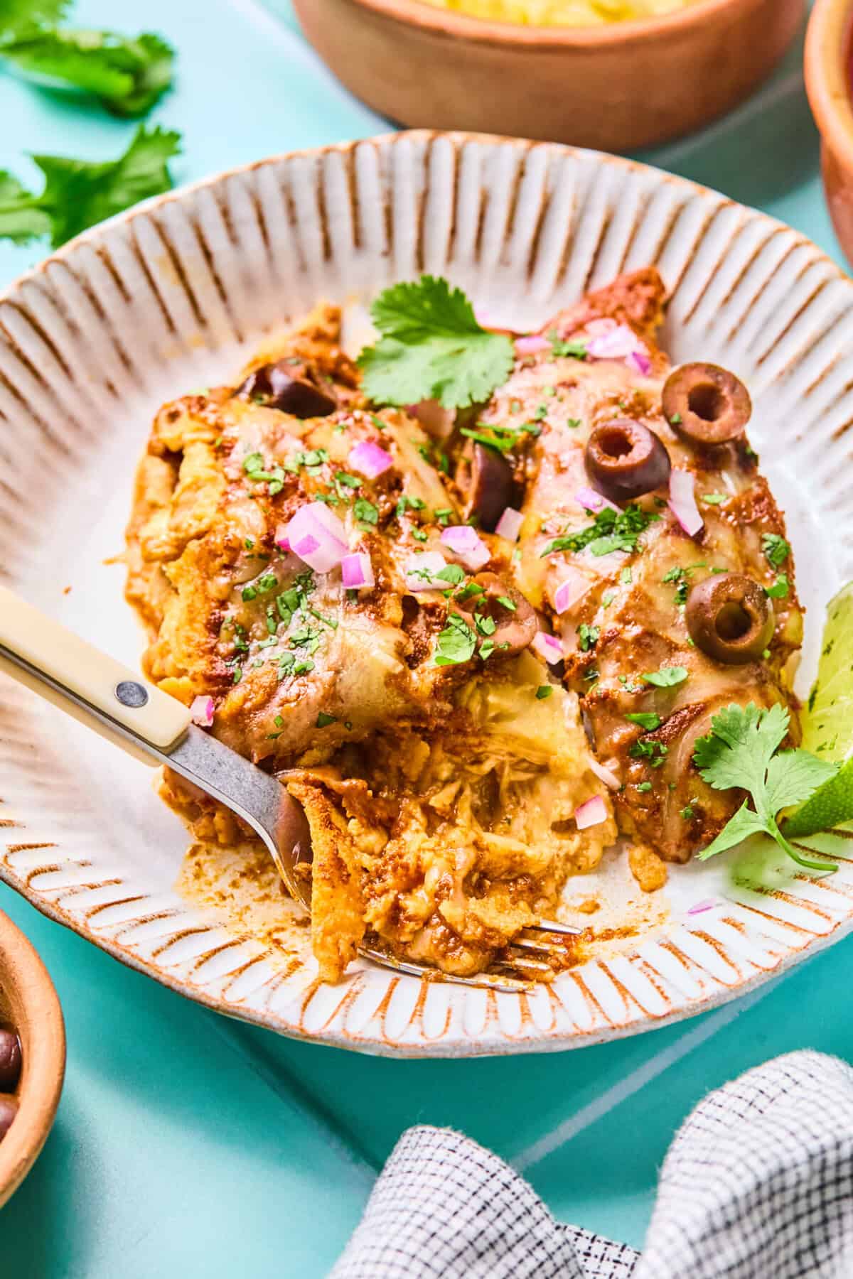 Two rotisserie chicken enchiladas topped with sauce, melted cheese, sliced black olives, chopped red onions, and cilantro on a ceramic plate, with a fork in one enchilada and a sprig of cilantro on the side.