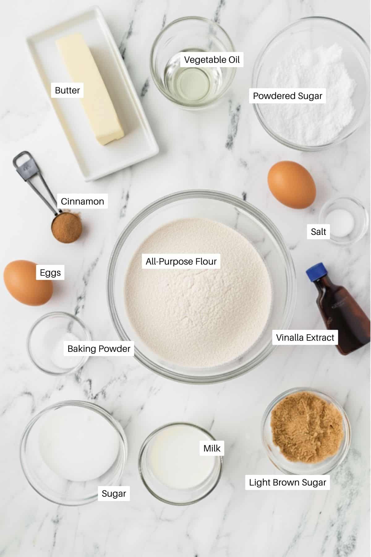 A marble countertop displays labeled baking ingredients for Coffee Cake Muffins: butter, eggs, cinnamon, vegetable oil, powdered sugar, salt, vanilla extract, all-purpose flour, baking powder, milk, sugar, and light brown sugar in bowls and containers.