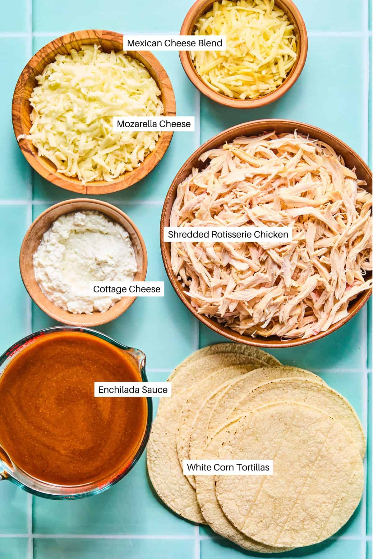 Bowls containing ingredients for rotisserie chicken enchiladas—Mexican cheese blend, mozzarella, shredded rotisserie chicken, cottage cheese, and enchilada sauce—along with white corn tortillas, are arranged on a turquoise tiled surface, each labeled.