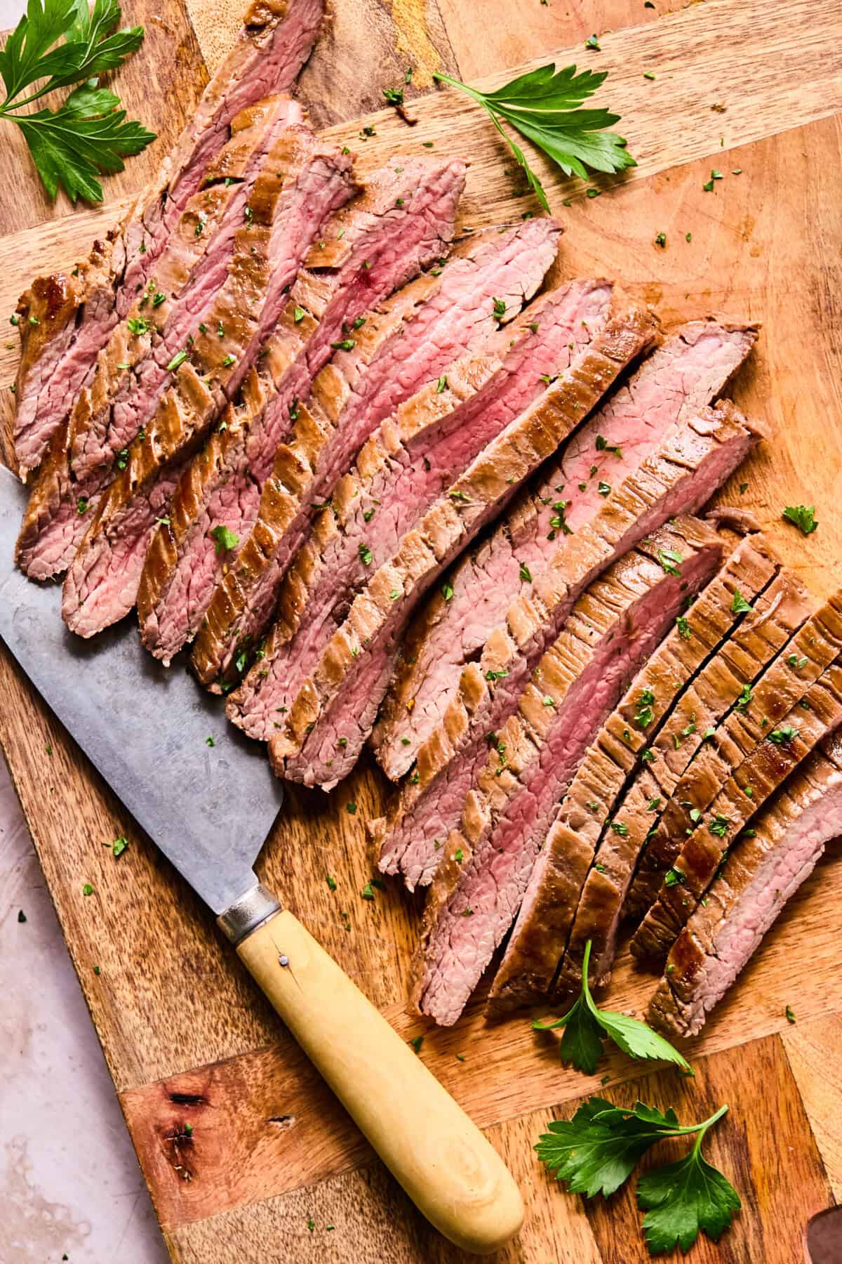 Sliced Air Fryer Flank Steak arranged neatly on a wooden cutting board, garnished with chopped herbs. A large knife with a wooden handle rests beside the meat, while fresh parsley leaves are scattered around.