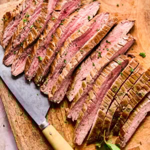 Sliced Air Fryer Flank Steak arranged neatly on a wooden cutting board, garnished with chopped herbs. A large knife with a wooden handle rests beside the meat, while fresh parsley leaves are scattered around.