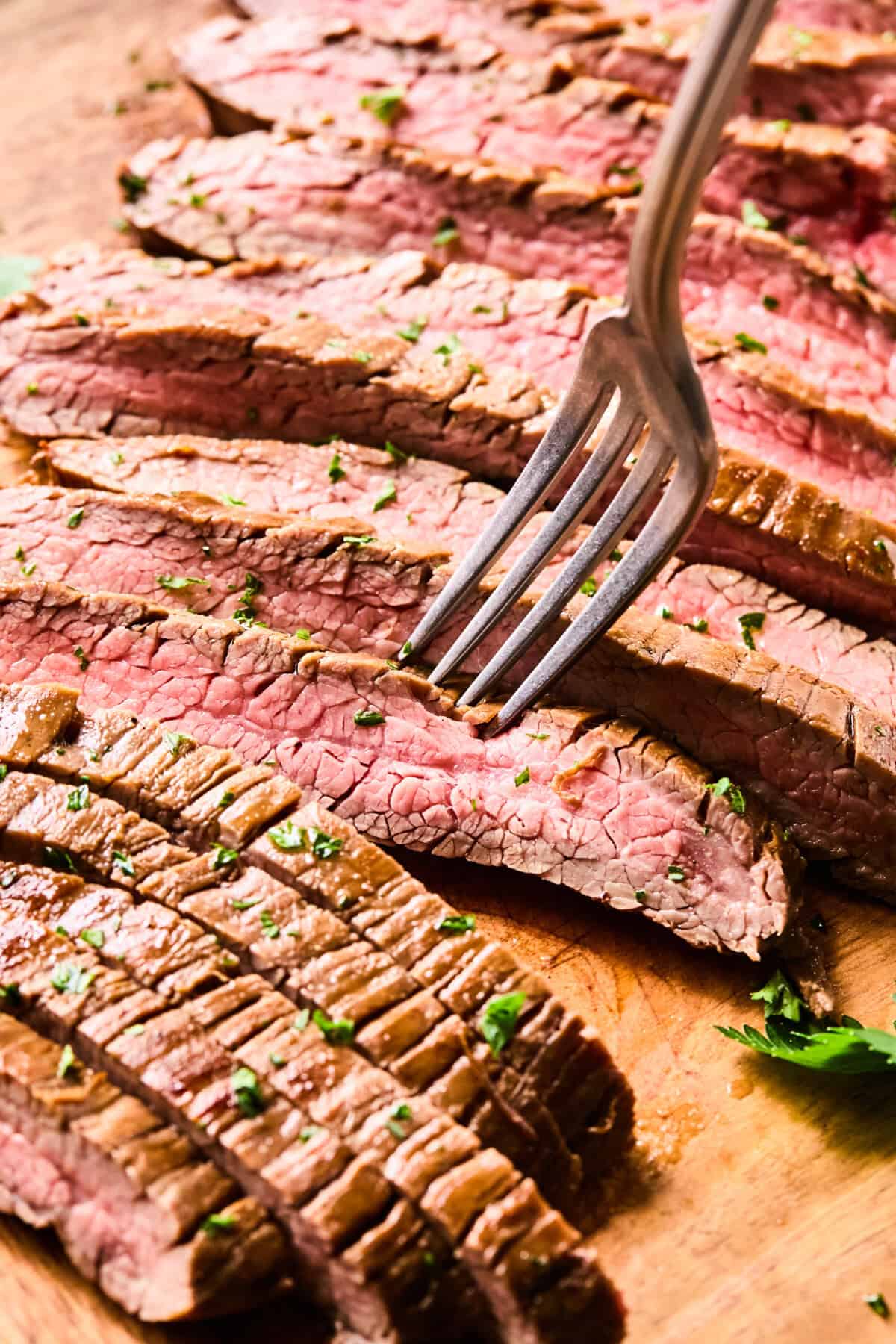 Air Fryer Flank Steak, sliced medium-rare and garnished with herbs, rests on a wooden board, with a fork piercing one juicy slice.