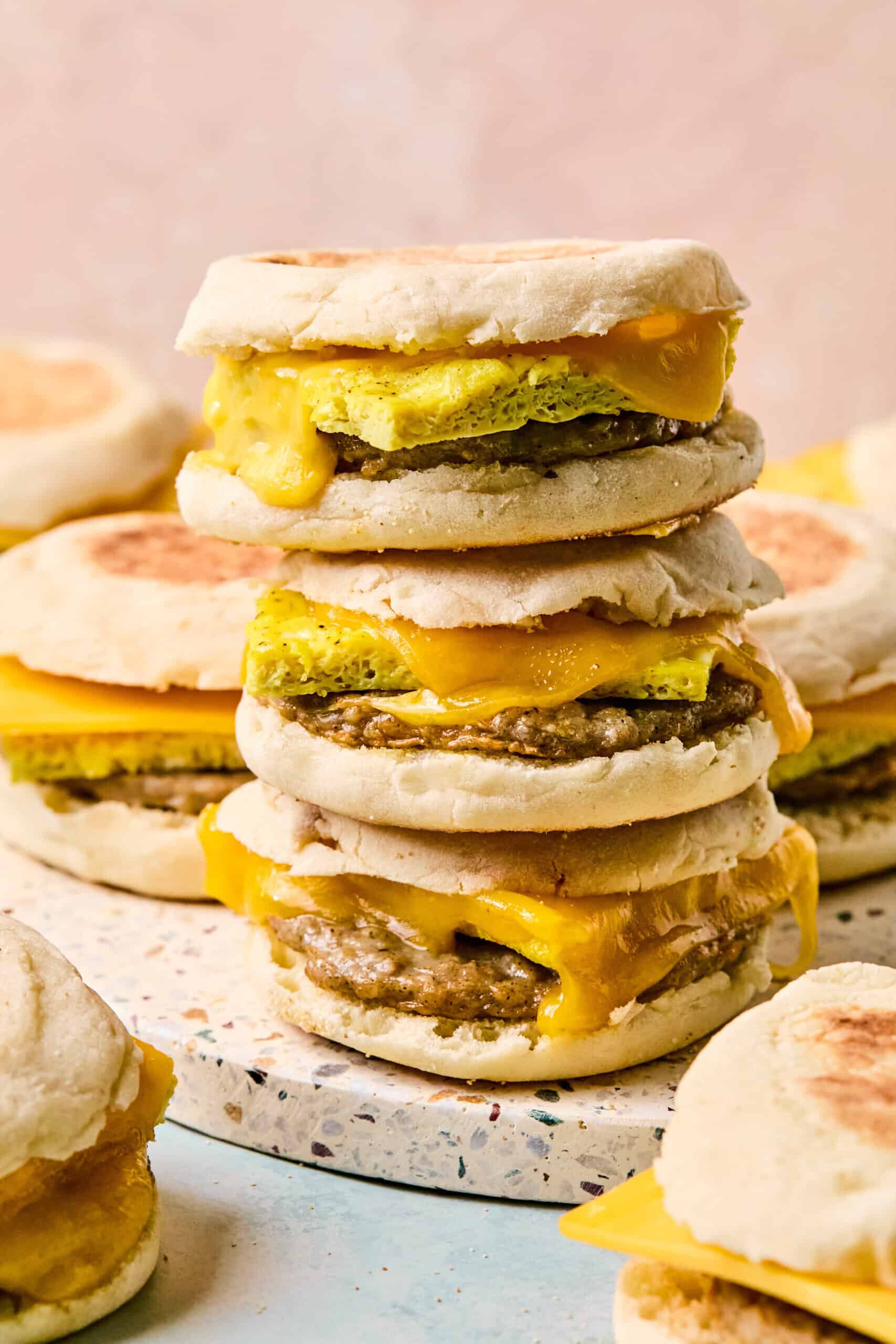 A close-up of breakfast sandwiches on English muffin, with eggs, sausage patties, and melted cheddar cheese stacked high on a speckled surface, more tempting sandwiches in the background.