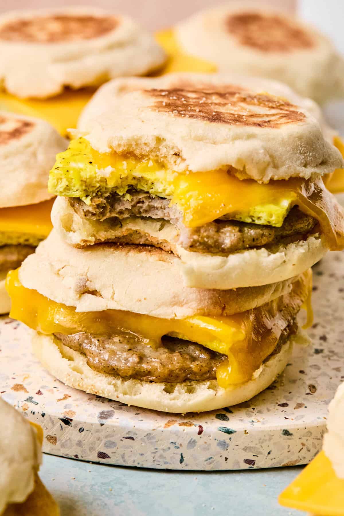 Two breakfast sandwiches on English muffin with sausage, egg, and melted cheddar cheese are stacked on a surface. The top sandwich has a bite taken out, revealing the filling. More breakfast sandwiches are visible in the background.