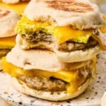 Two breakfast sandwiches on English muffin with sausage, egg, and melted cheddar cheese are stacked on a surface. The top sandwich has a bite taken out, revealing the filling. More breakfast sandwiches are visible in the background.