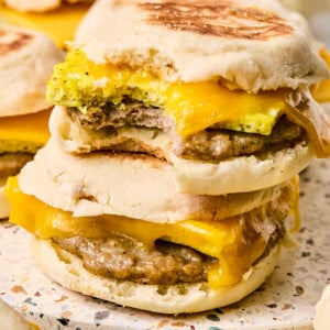 Two breakfast sandwiches on English muffin with sausage, egg, and melted cheddar cheese are stacked on a surface. The top sandwich has a bite taken out, revealing the filling. More breakfast sandwiches are visible in the background.
