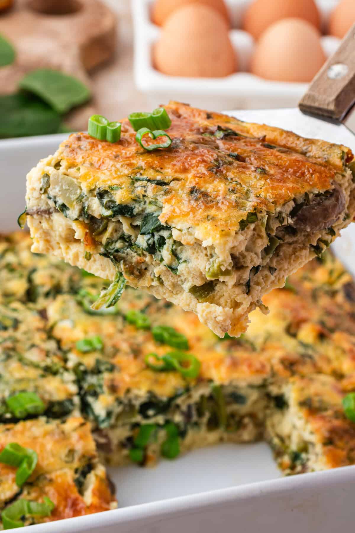 A close-up of a golden brown, fluffy cottage cheese egg casserole with spinach and green onions being lifted from a dish, with whole eggs and fresh spinach in the background.