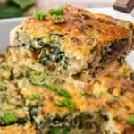 A close-up of a golden brown, fluffy cottage cheese egg casserole with spinach and green onions being lifted from a dish, with whole eggs and fresh spinach in the background.