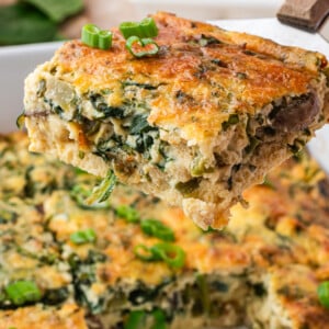 A close-up of a golden brown, fluffy cottage cheese egg casserole with spinach and green onions being lifted from a dish, with whole eggs and fresh spinach in the background.