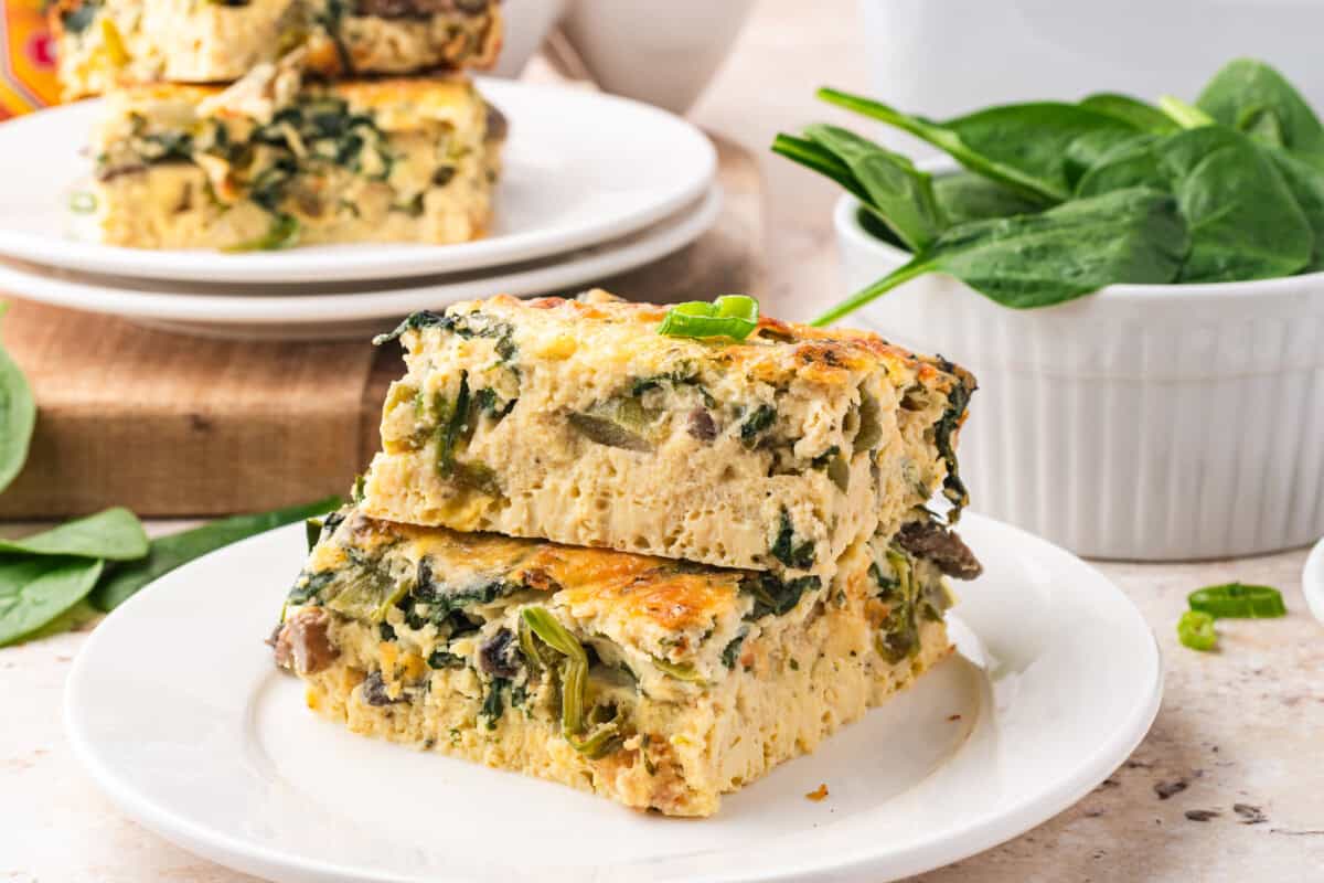 Two square slices of vegetable frittata with spinach and mushrooms, reminiscent of a cottage cheese egg casserole, are stacked on a white plate, with fresh spinach leaves and more frittata seen in the background.