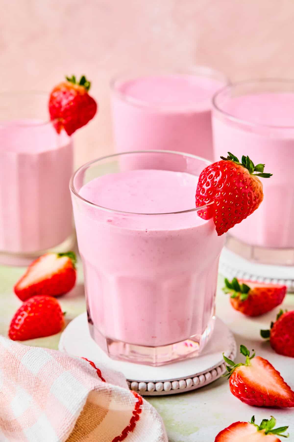 Three glasses of pink strawberry protein shake, each garnished with a fresh strawberry, sit on coasters. Sliced strawberries and a striped cloth are nearby on a light surface with a pastel background.