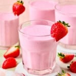 Three glasses of pink strawberry protein shake, each garnished with a fresh strawberry, sit on coasters. Sliced strawberries and a striped cloth are nearby on a light surface with a pastel background.