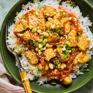 A bowl of white rice topped with sweet chili chicken, red bell peppers, chopped green onions, and crushed peanuts, served with a fork on the side.