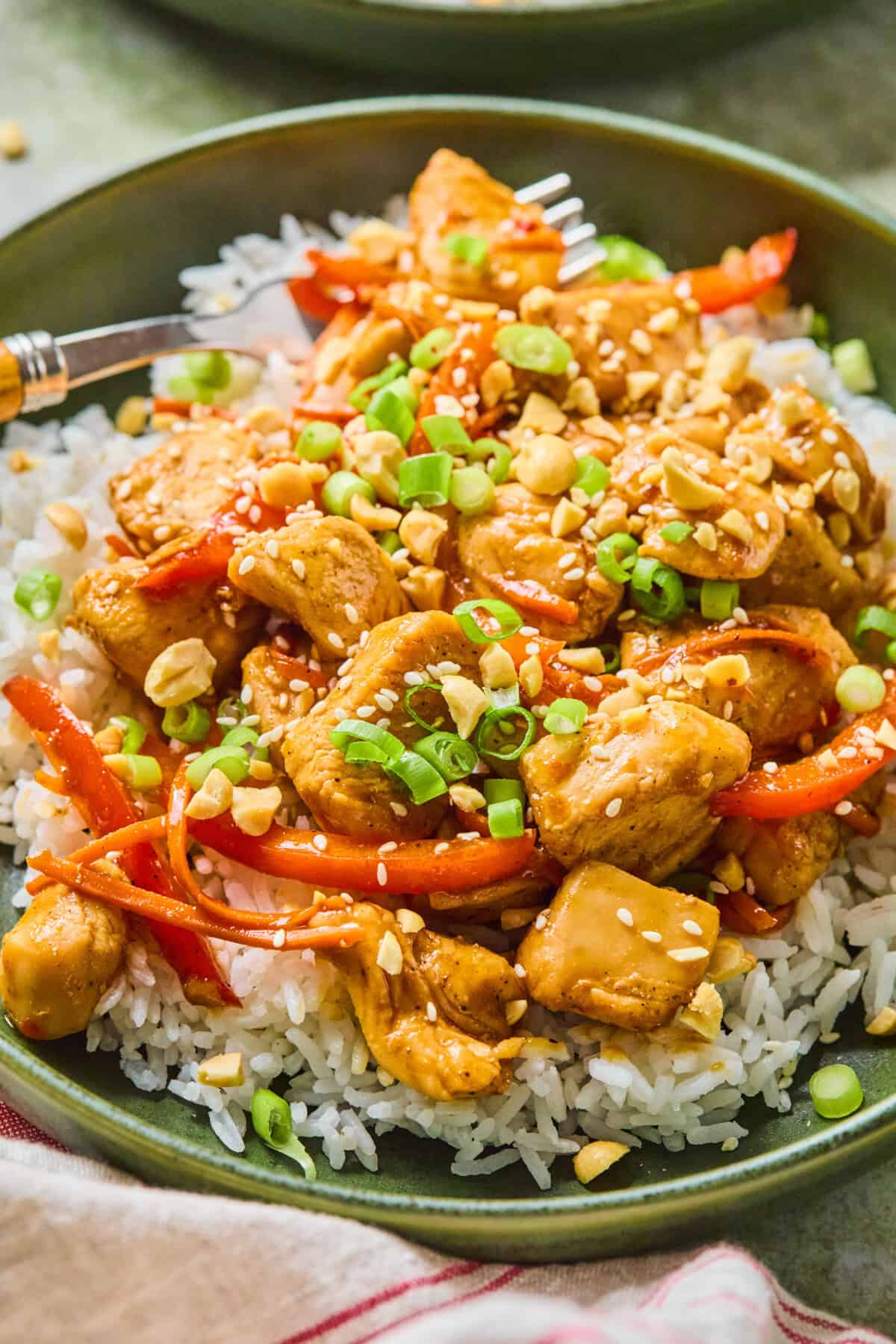 A bowl of white rice topped with sweet chili chicken, red bell peppers, chopped green onions, peanuts, and sesame seeds, with a fork resting on the side.