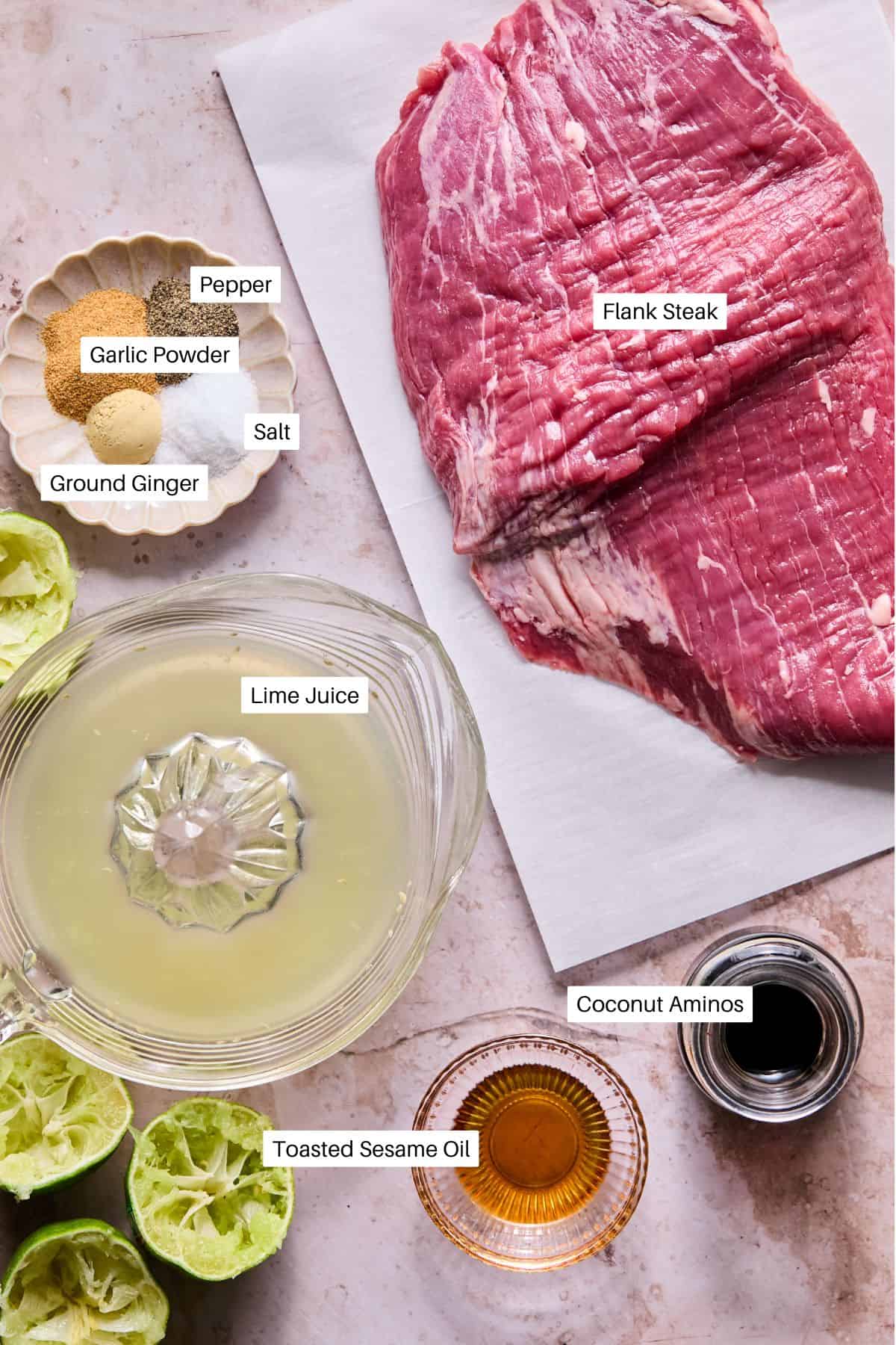 Raw flank steak on a cutting board, ready to become Air Fryer Flank Steak, is surrounded by bowls of pepper, garlic powder, salt, ground ginger, coconut aminos, toasted sesame oil, and a pitcher of lime juice with squeezed lime halves nearby. Each item is labeled.