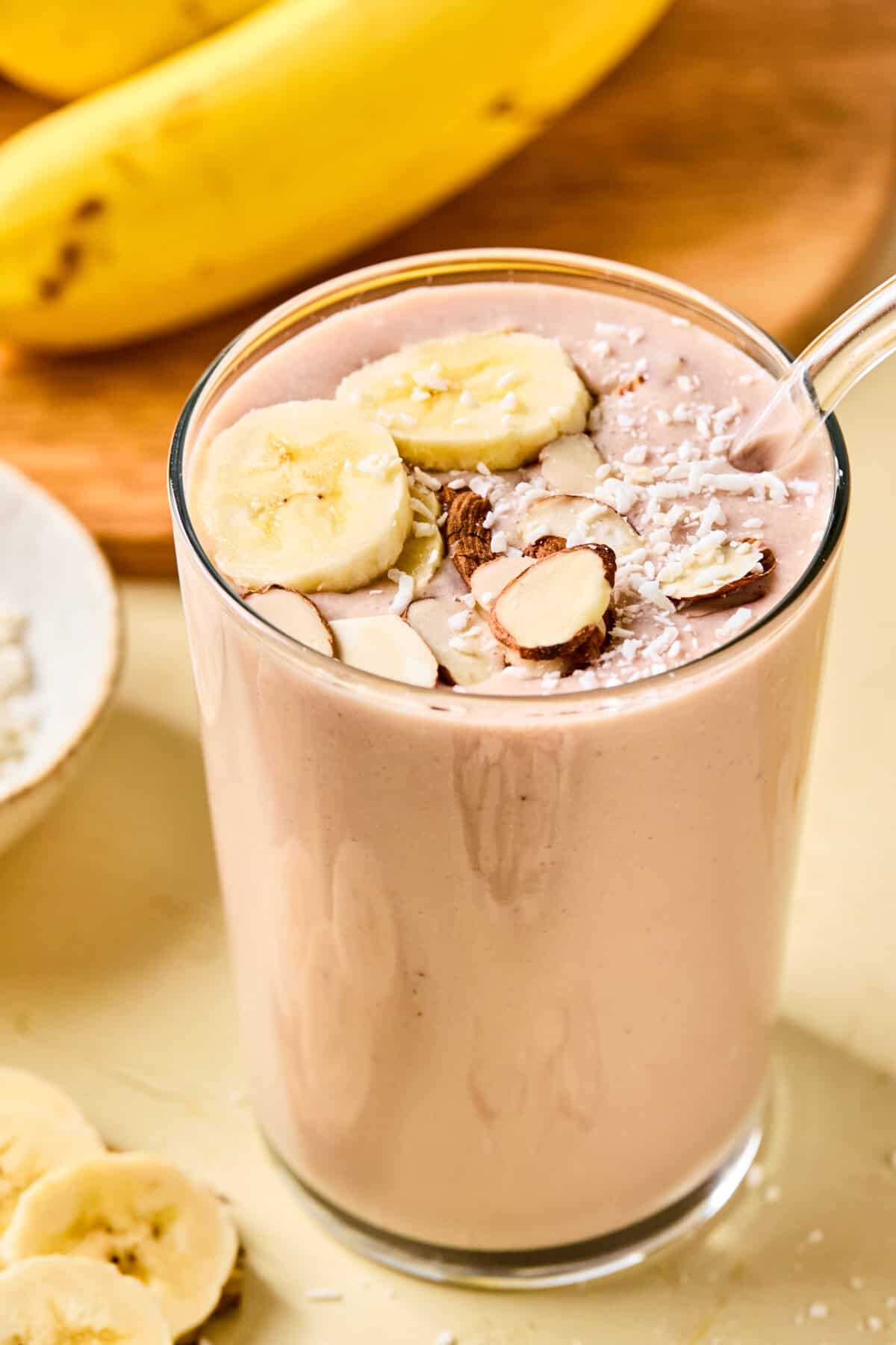 A glass of banana protein shake topped with banana slices, almond slivers, and shredded coconut sits on a light surface. Whole bananas and sliced pieces are visible in the background.