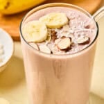 A glass of banana protein shake topped with banana slices, almond slivers, and shredded coconut sits on a light surface. Whole bananas and sliced pieces are visible in the background.