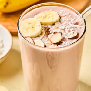 A glass of banana protein shake topped with banana slices, almond slivers, and shredded coconut sits on a light surface. Whole bananas and sliced pieces are visible in the background.