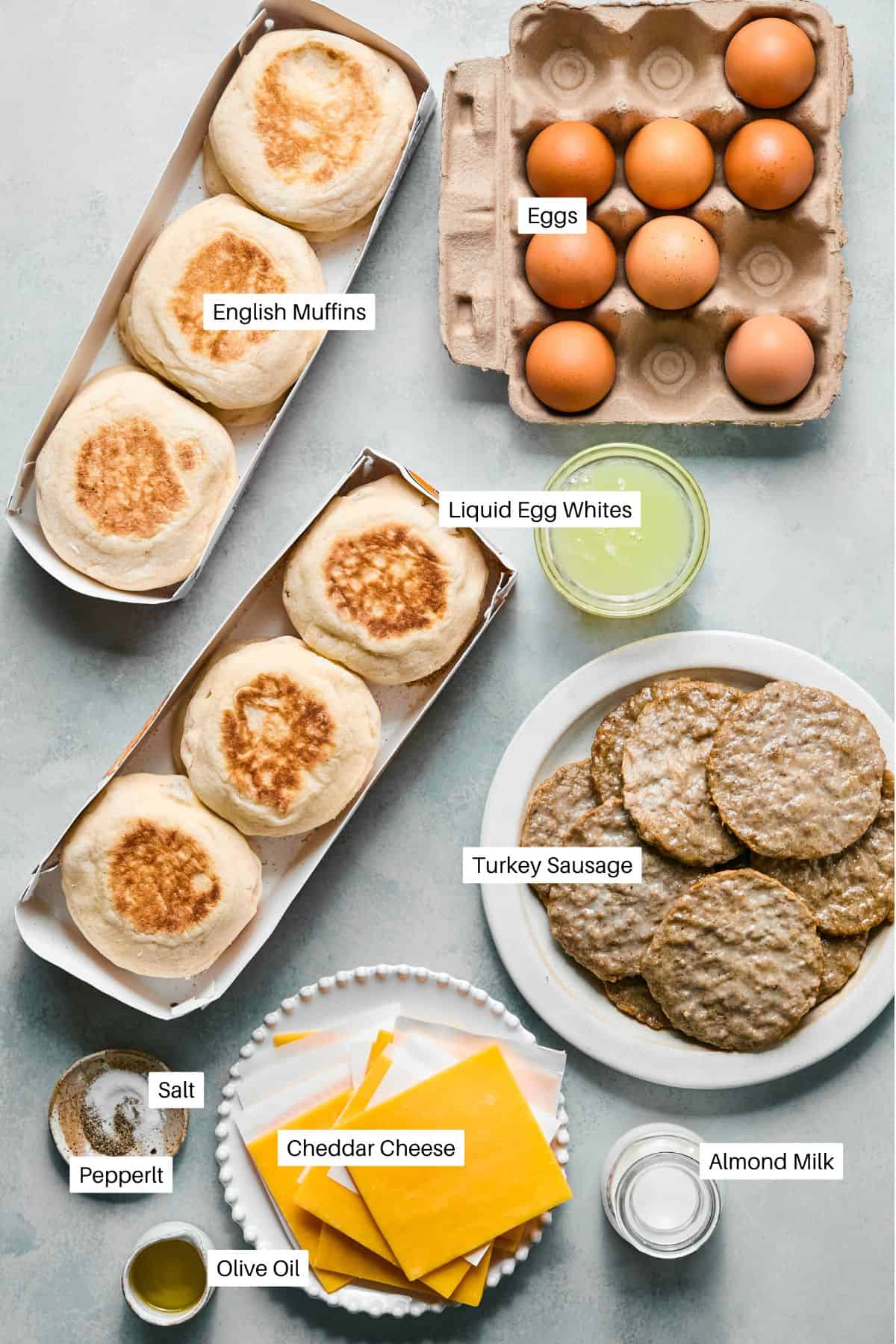 Top-down view of labeled ingredients for breakfast sandwiches on English muffin: eggs, liquid egg whites, turkey sausage patties, cheddar cheese slices, almond milk, olive oil, salt, and pepper on a light surface.